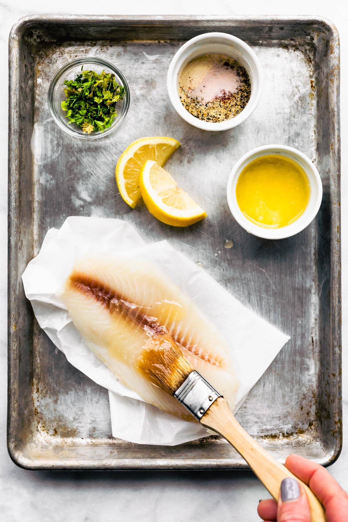 With ingredients on a baking sheet a hand is spreading melted butter onto the raw fish fillet.