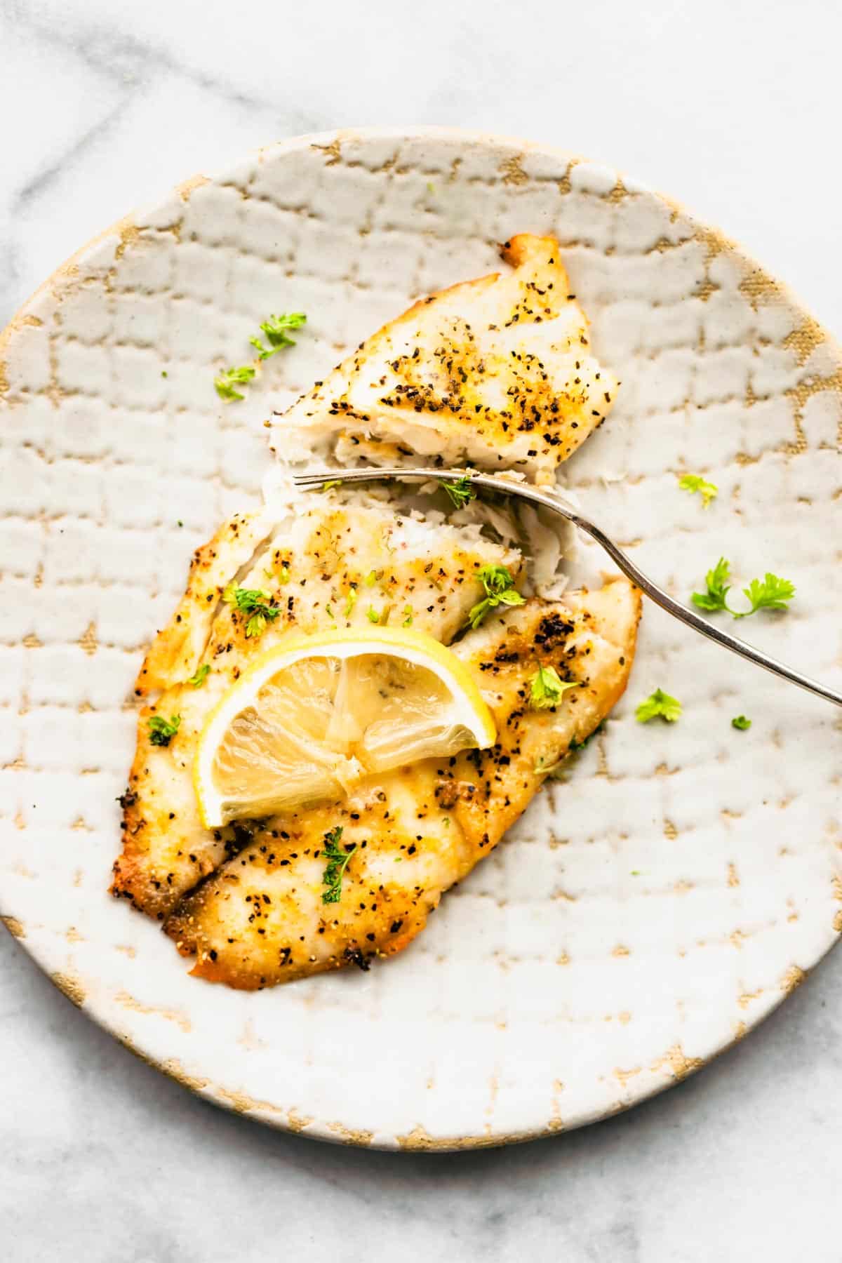 Cooked Tilapia topped with lemon on a plate being cut up by a fork