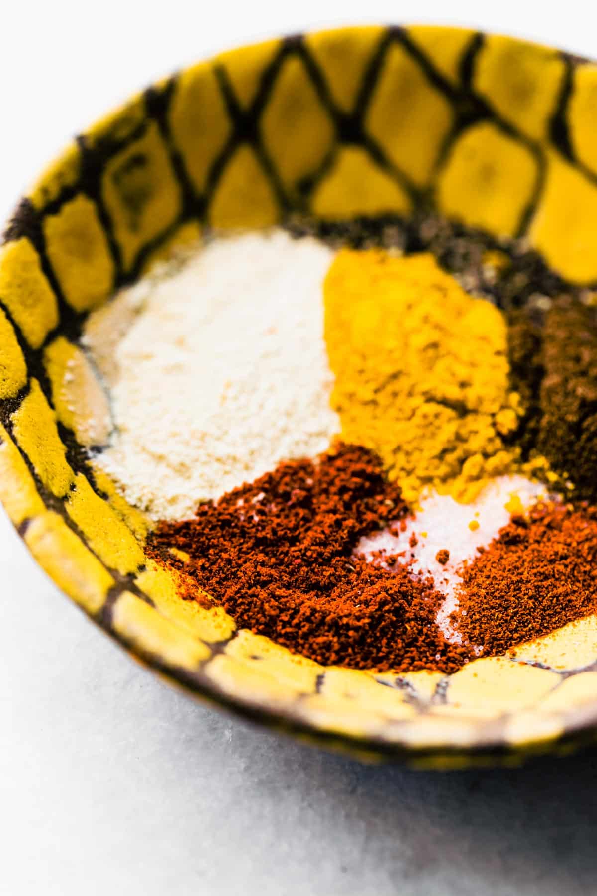 Close-up shot of various spices (salt, turmeric, allspice, cayenne, paprika & black pepper)  in a yellow bowl.