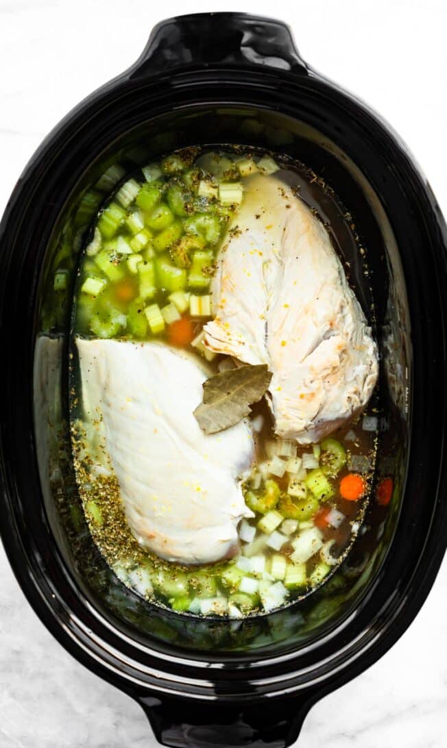 two chicken breasts in a slow cooker with celery, onions, carrots, broth, and a bay leaf