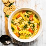 a bowl of gluten-free chicken noodle soup on a platter with a spoon and gluten-free crackers