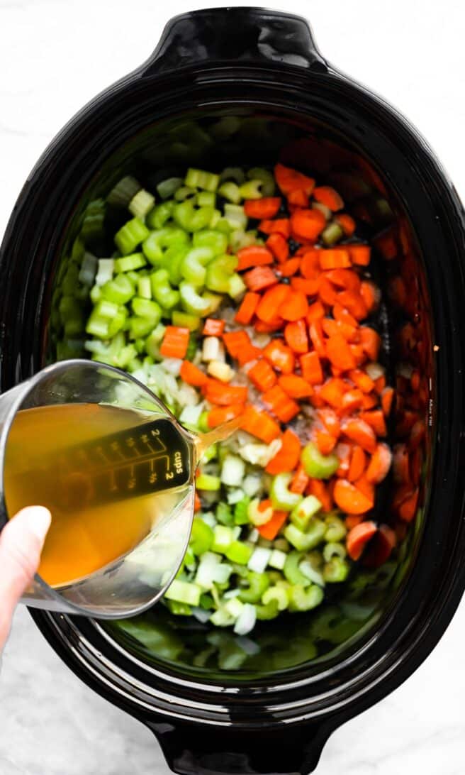 broth being poured over celery, onions, and carrots in a slow cooker