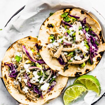 2 pork tacos with red cabbage and cilantro on top. Lime on side. Sitting on a white plate