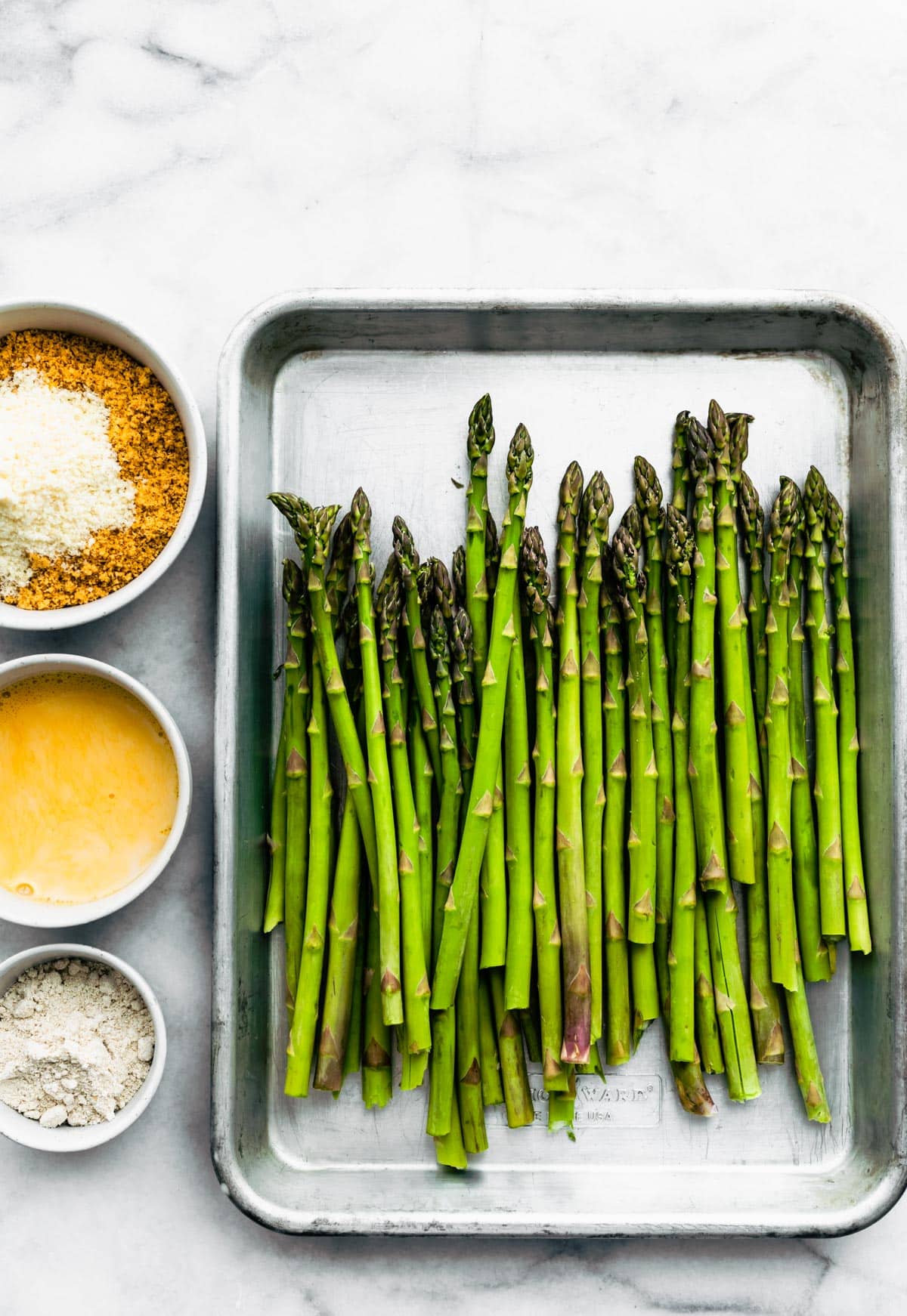 Spears of asparagus in a baking dish, a bowl of gluten-free breadcrumbs, a bowl of whisked eggs, and Parmesan.