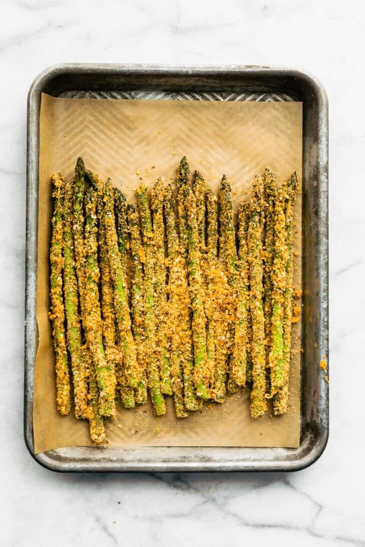 Uncooked parmesan panko air fryer asparagus fries on a parchment paper-lined baking sheet.