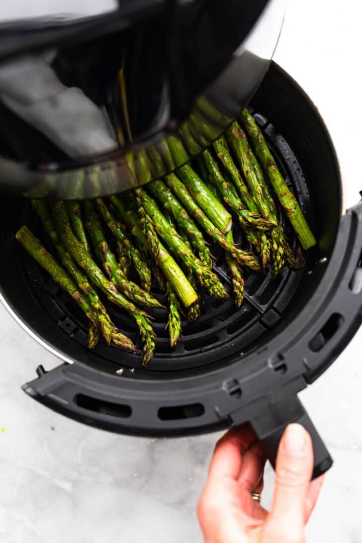 An air fryer basket full of cooked asparagus spears being pulled out of the air fryer.