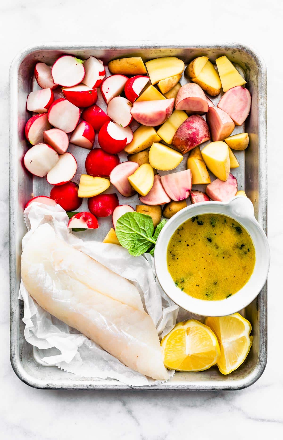 Easy Baked Fish and Vegetables ingredients on a baking sheet - fish fillet, lemons, potatoes, radishes, and a mustard sauce.