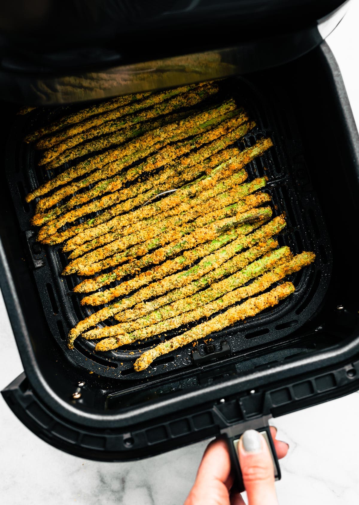A hand holding an air fryer basket full of parmesan coated asparagus fries.