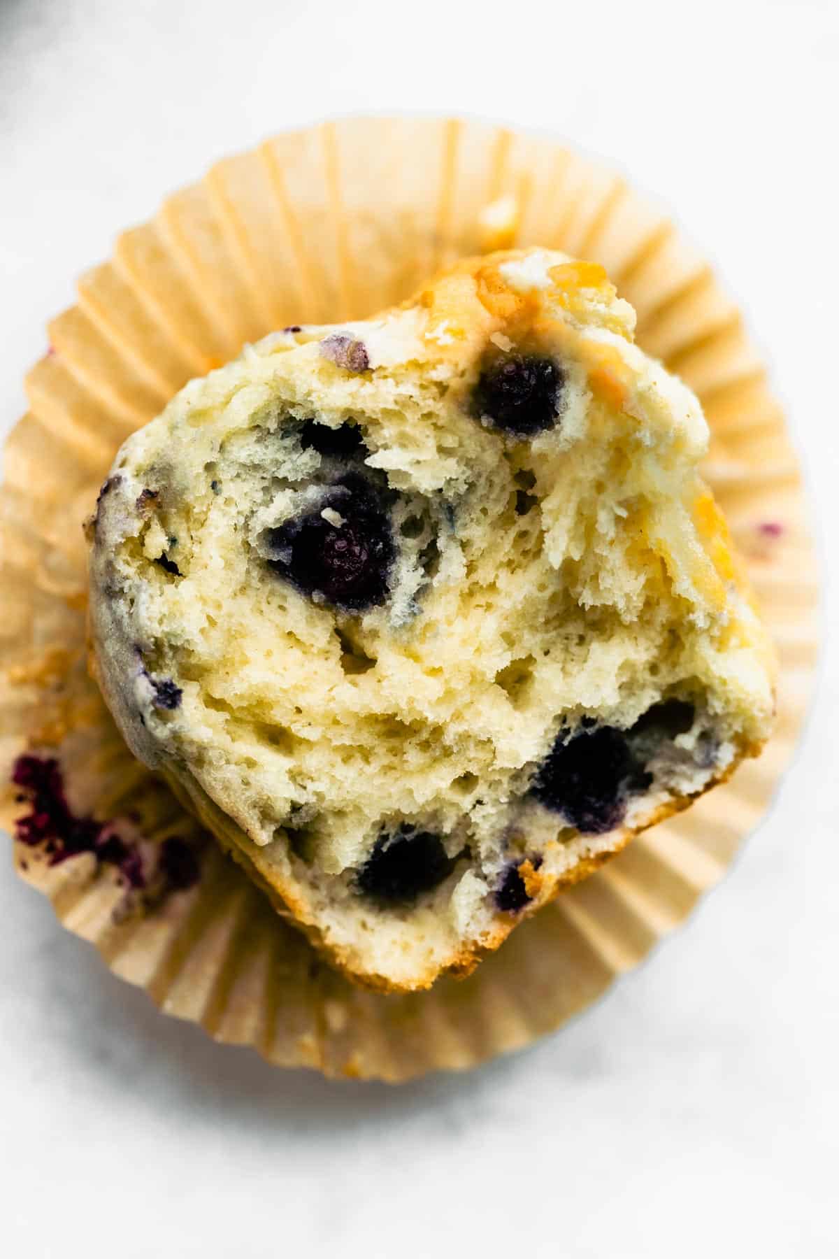 A gluten free blueberry muffin on top of a muffin wrapper with a bite missing.
