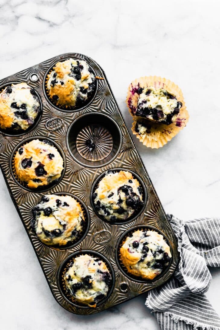A muffin tin full of gluten free blueberry muffins with one muffin missing and off to the side on top of a wrapper with a bite taken out.