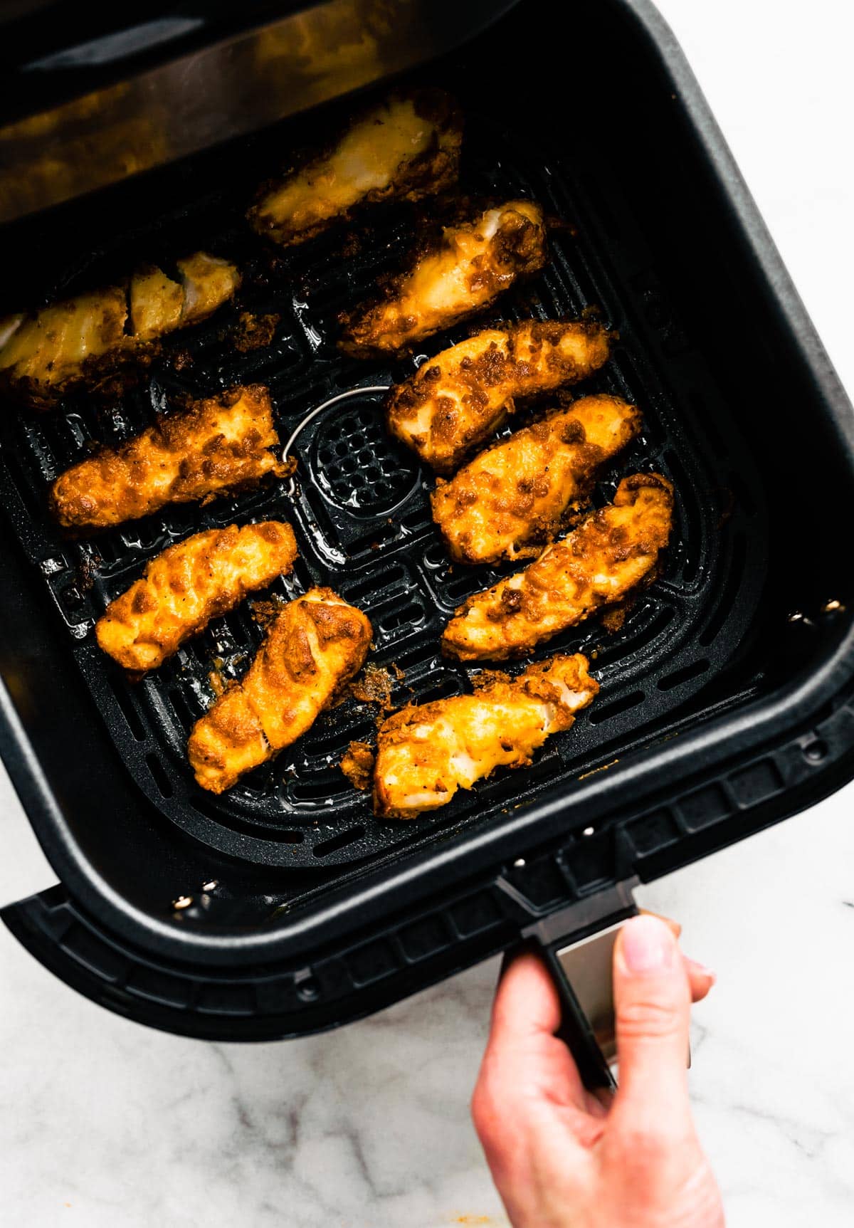 A hand pulling out an air fryer basket full of pieces of gluten-free beer-battered fish.