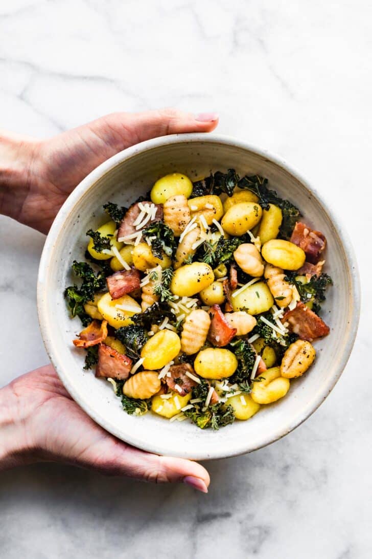 Two hands holding a bowl of gluten-free gnocchi with bacon, kale, and Parmesan cheese.