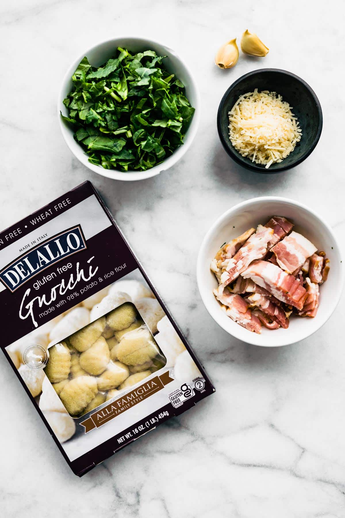 Overhead image of a bowl of kale, two garlic cloves, shredded Parmesan, chopped bacon, and a box of Delallo gluten-free gnocchi.