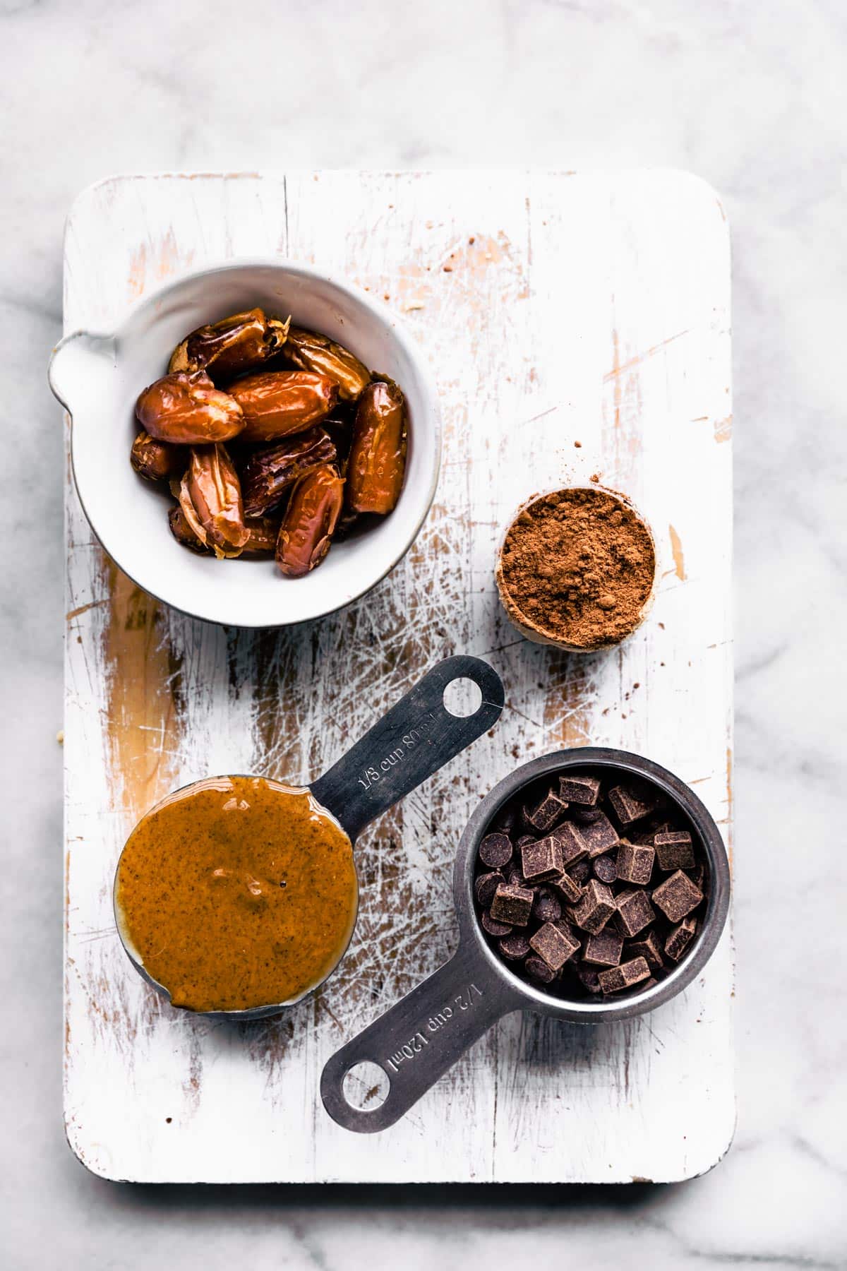 A bowl of dates, cocoa powder, sunflower seed butter, and chocolate chunks on a white wood cutting board.