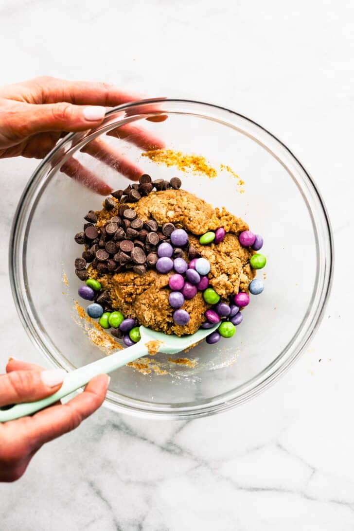 Two hands using a rubber spatula to fold chocolate chips and chocolate candies into gluten free cookie bar batter.