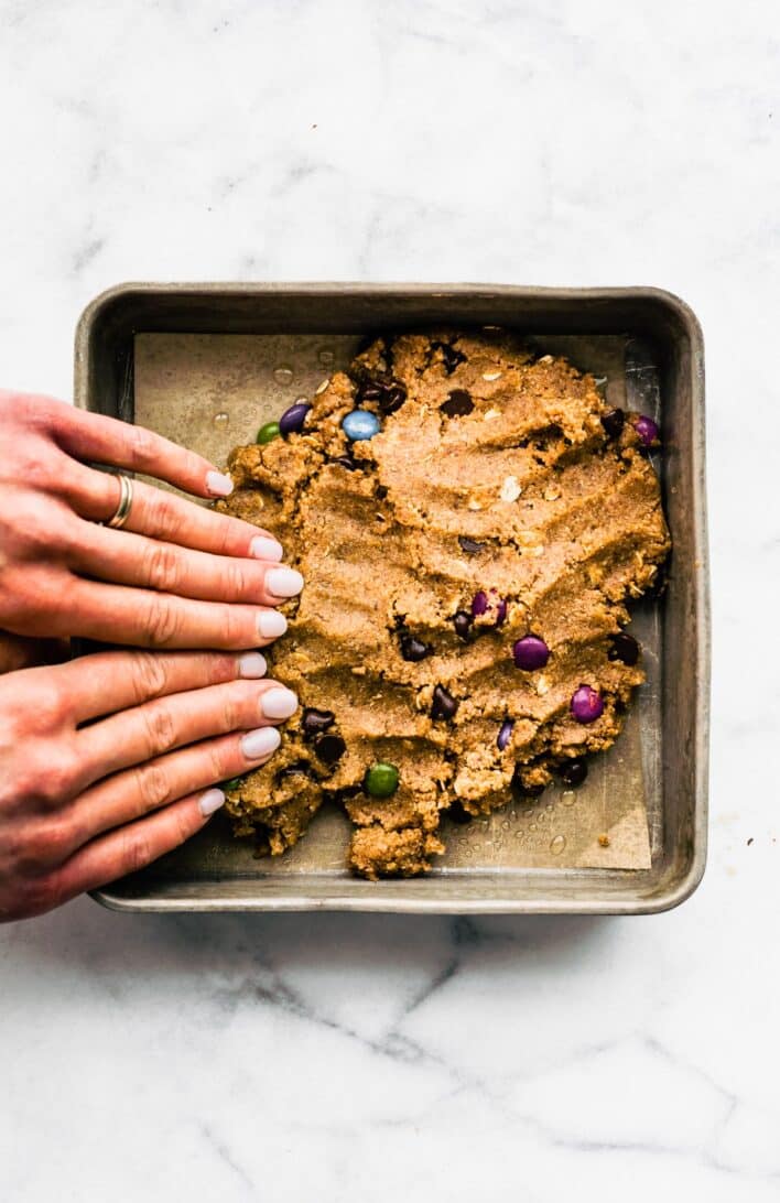 Two hands pushing gluten free cookie bar batter into a square baking pan lined with parchment paper.