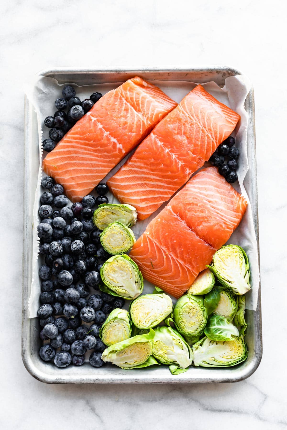 Overhead image of a sheet pan with three salmon filets, fresh blueberries, and Brussels sprouts.