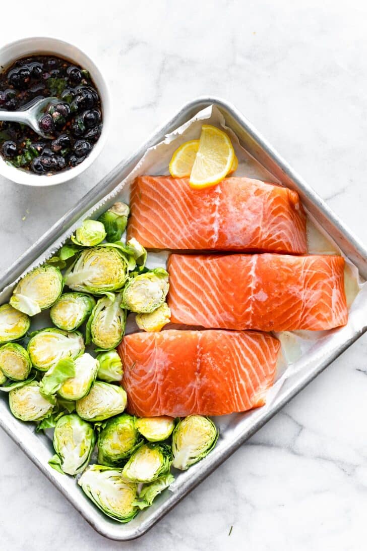 A sheet pan with three raw salmon filets and a side bowl of a blueberry mixture and Brussels sprouts.
