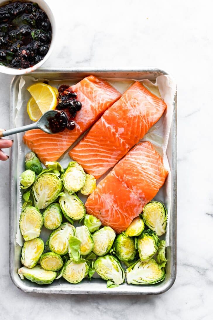 A sheet pan with three salmon filets and Brussels sprouts with a hand spooning a blueberry mixture on top of the salmon.