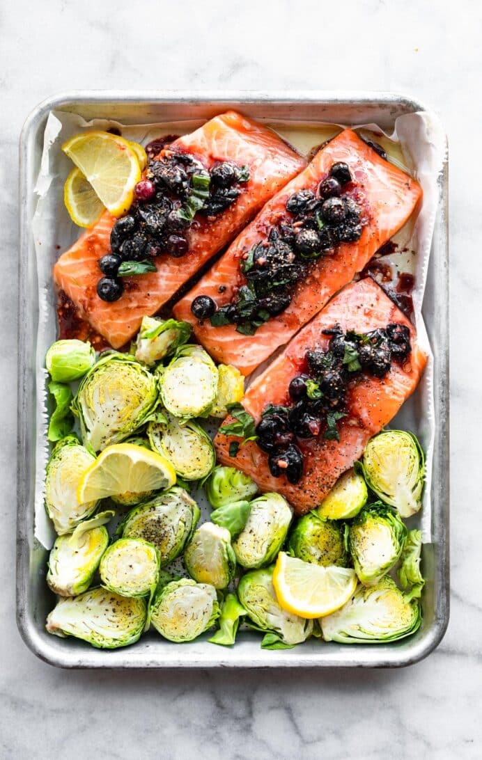 Overhead image of a baking dish with three salmon filets topped with blueberry compote and Brussels sprouts next to them.