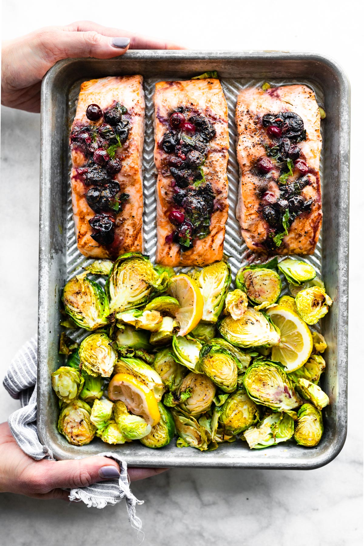 A sheet pan with three salmon filets topped with a blueberry mixture and Brussels sprouts.