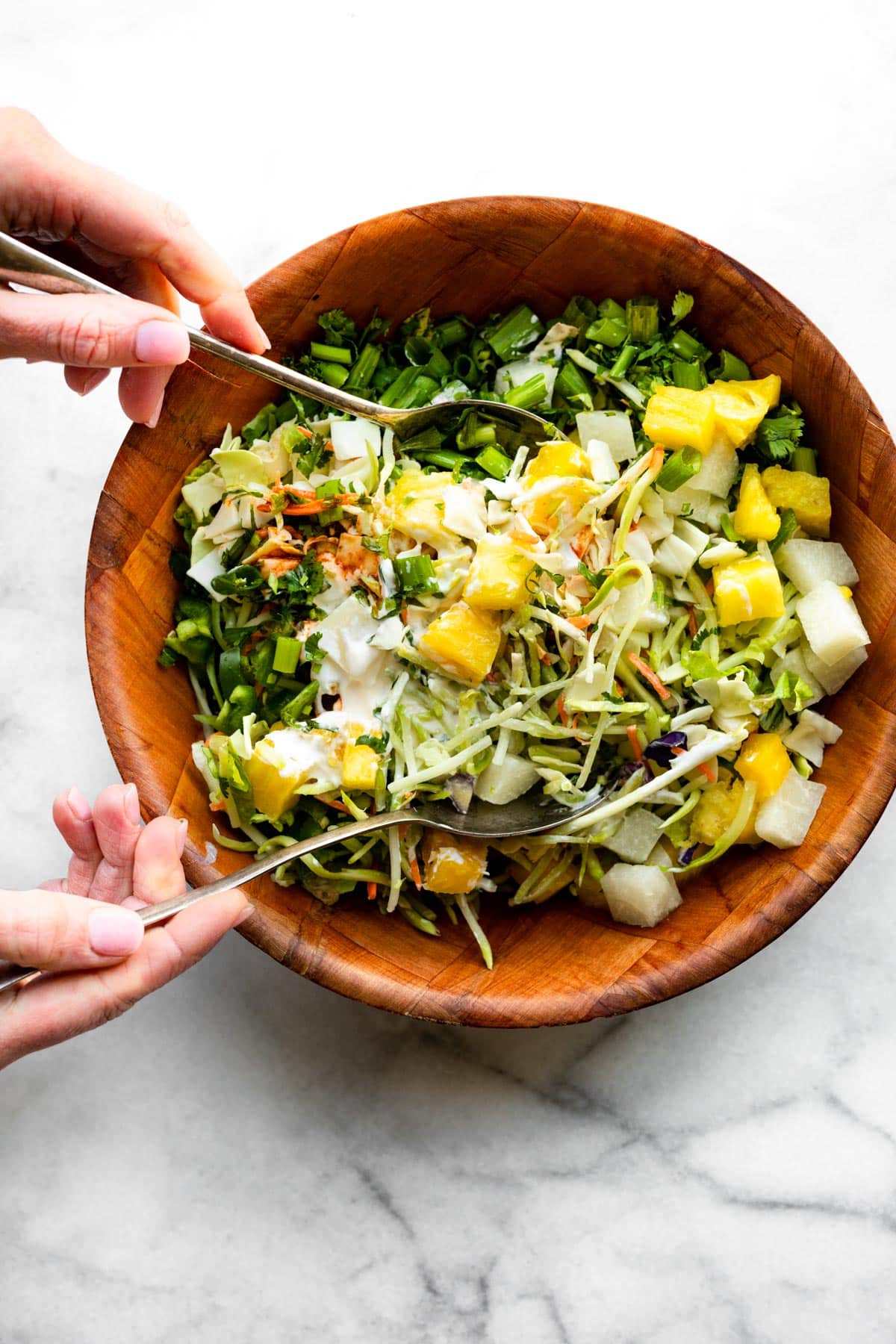 Two hands tossing pineapple coleslaw together in a wooden bowl.