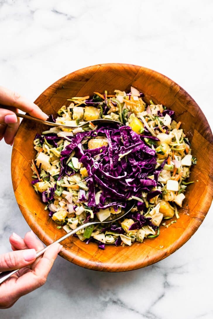 Two hands using serving spoons to fold extra red cabbage into a bowl of pineapple coleslaw.