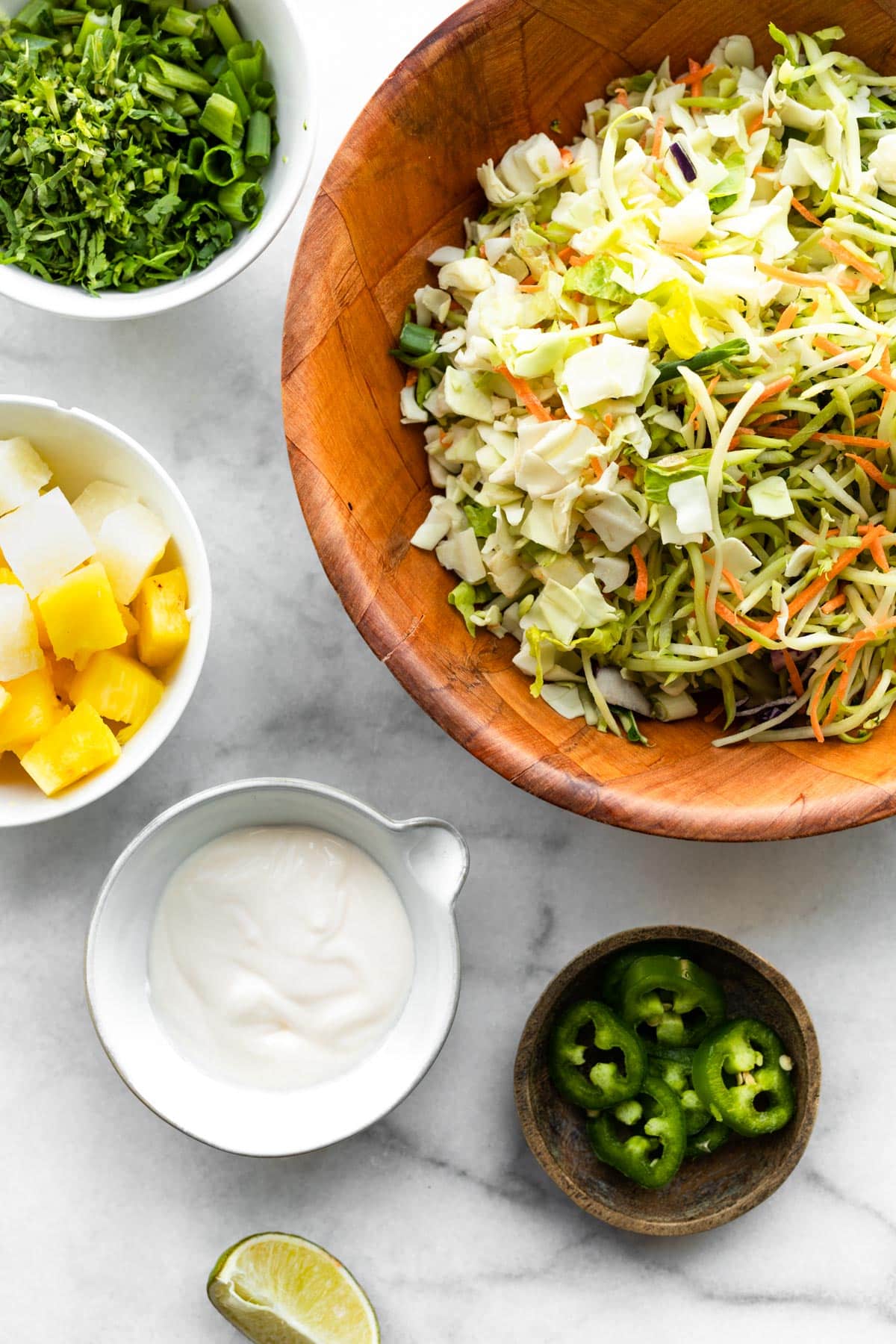 Overhead image of bowls of shredded cabbage and broccoli slaw, scallions, pineapple chunks, mayo, jalapeños, and a lime wedge.