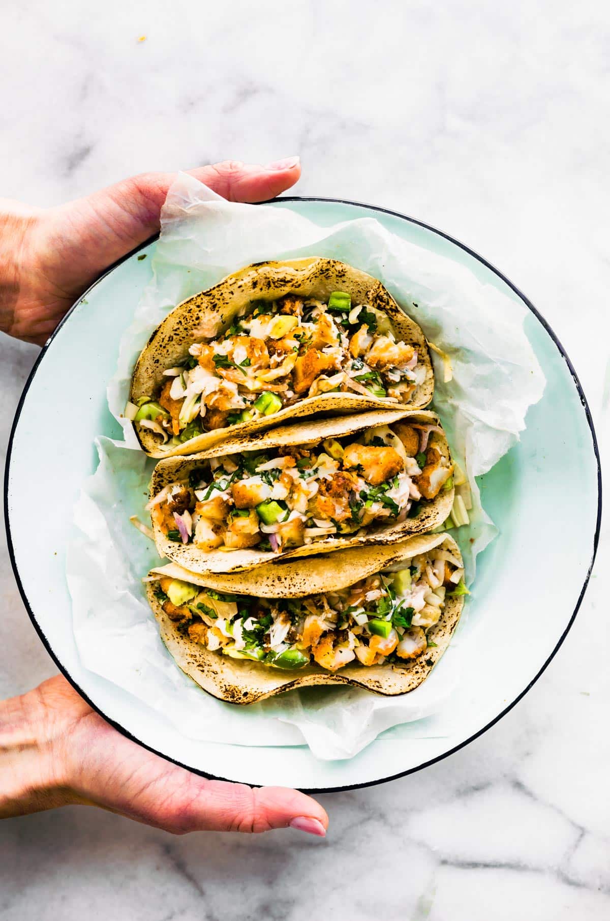 Two hands holding a bowl with three gluten-free air fryer fish tacos topped with pineapple slaw.
