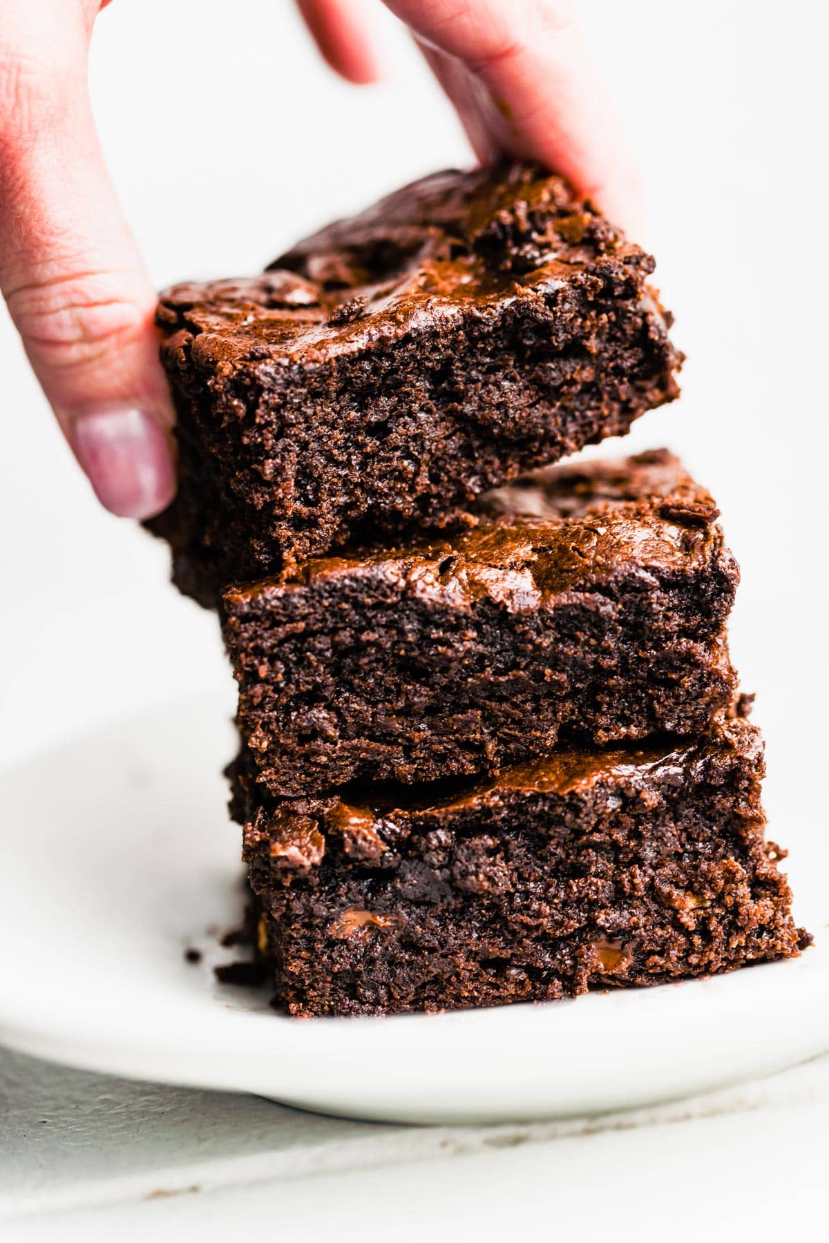 A hand grabbing a gluten free brownie from a stack of three brownies on top of each other on a plate.
