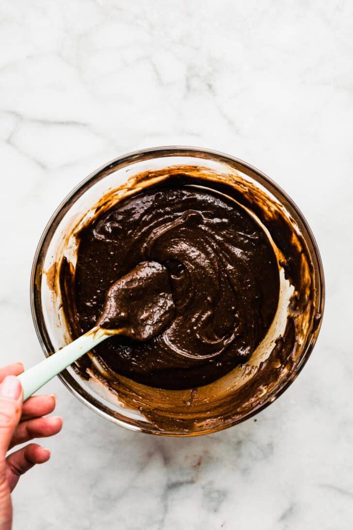 Overhead image of a glass bowl with gluten free brownie batter with a rubber spatula.
