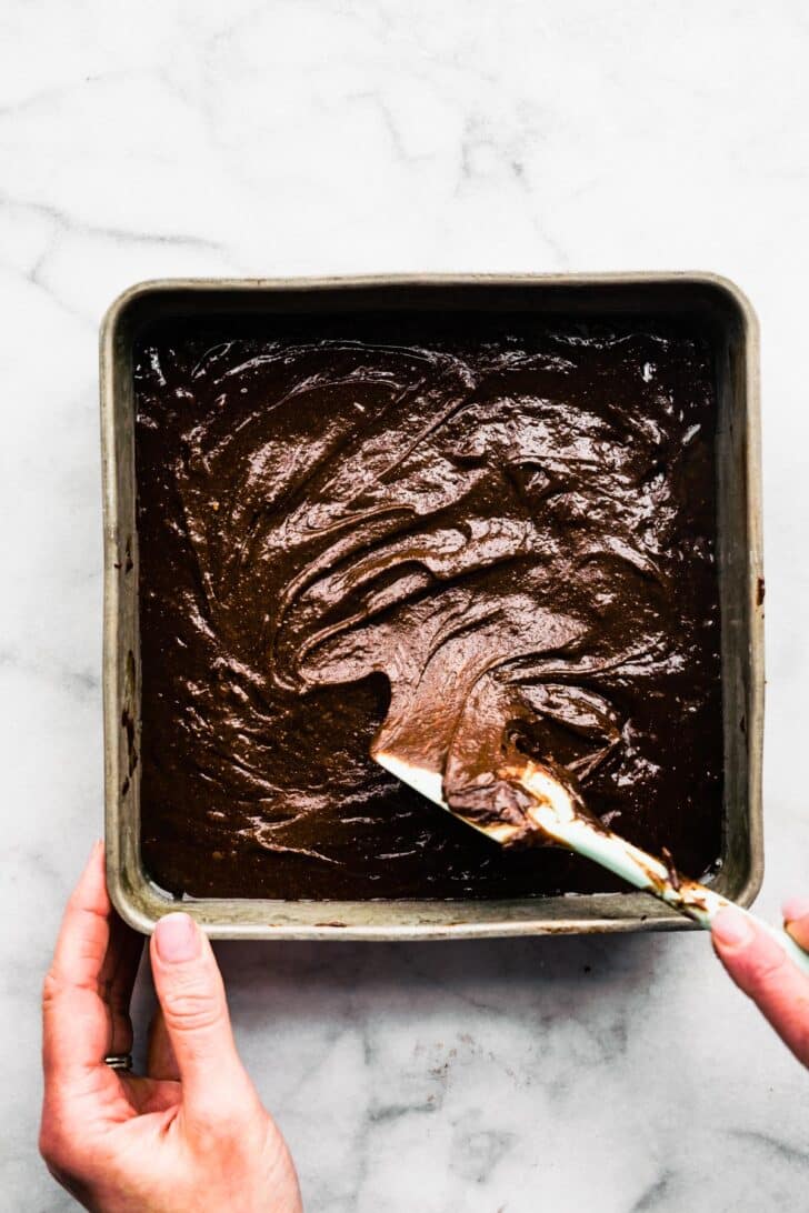 Gluten free brownie batter being spread out evenly in a square baking pan.