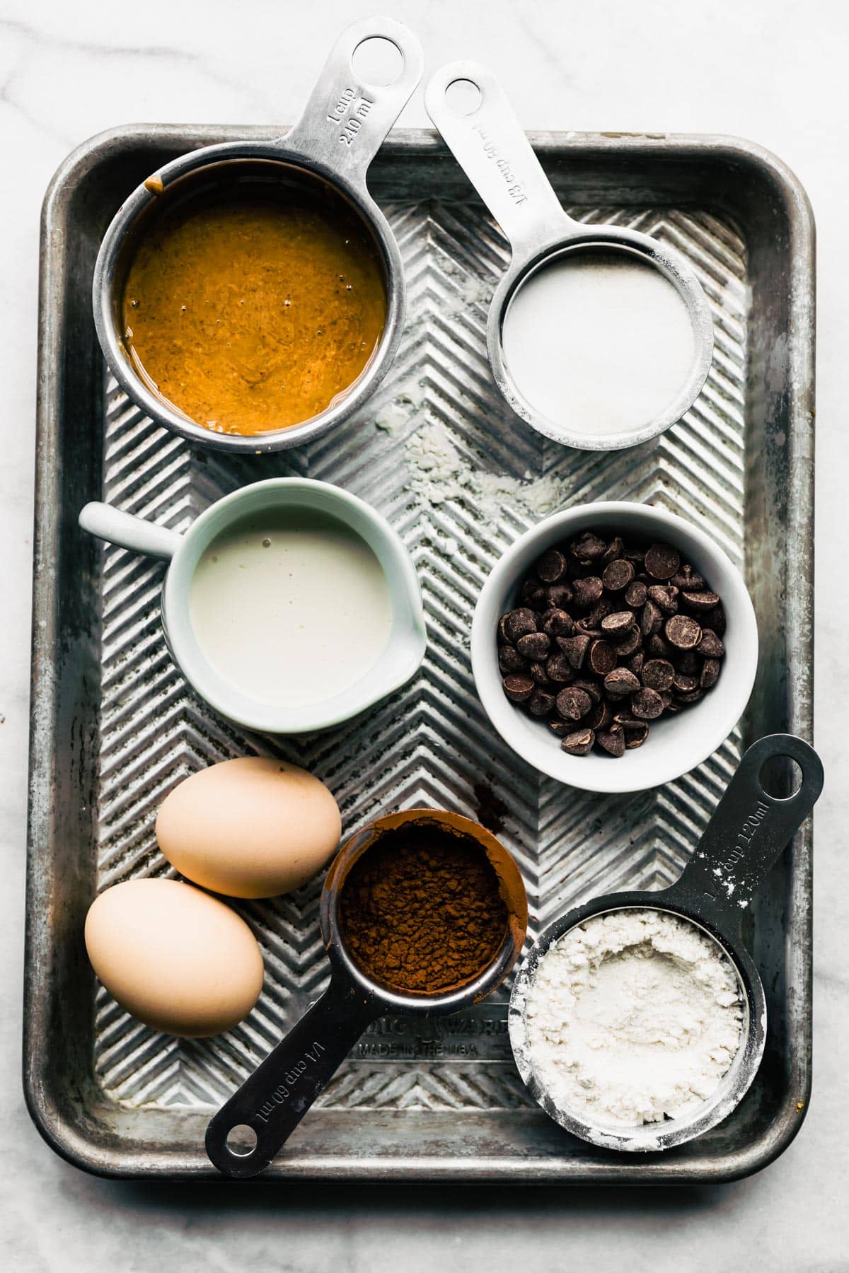 Overhead image of ingredients for gluten free brownies including nut butter, non-dairy milk, two eggs, cocoa powder, gluten-free flour, and chocolate chips.