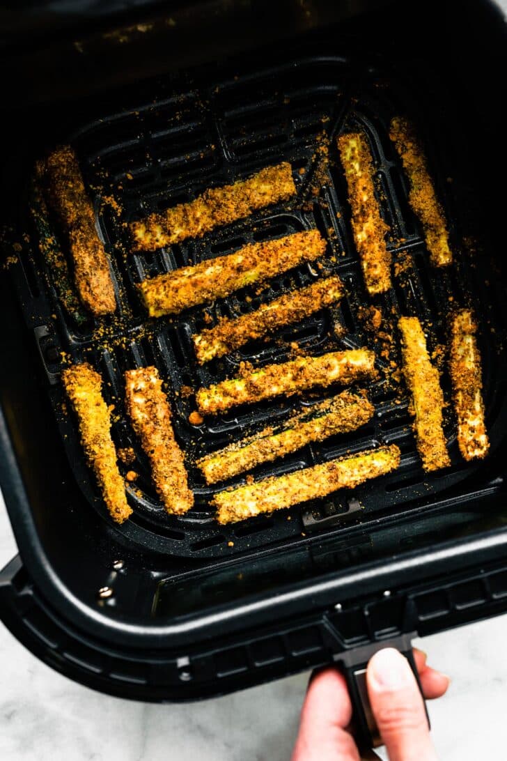 A hand holding an air fryer basket full of cooked air fryer zucchini fries.
