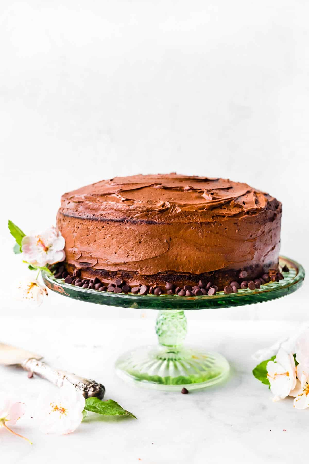 A gluten free chocolate cake iced with chocolate frosting on a cake stand surrounded by chocolate chips.