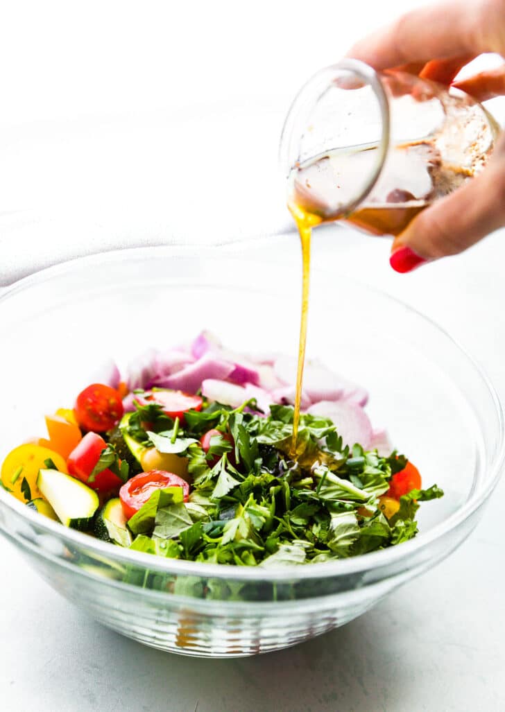 A hand pouring dressing over a bowl of tomato zucchini salad.