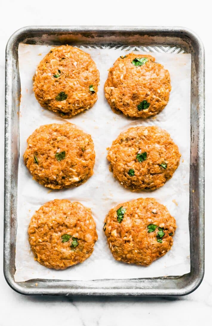 6 raw chicken burgers on a parchment paper-lined baking sheet.