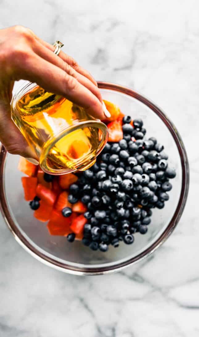 A hand pouring honey over strawberries and blueberries in a bowl.