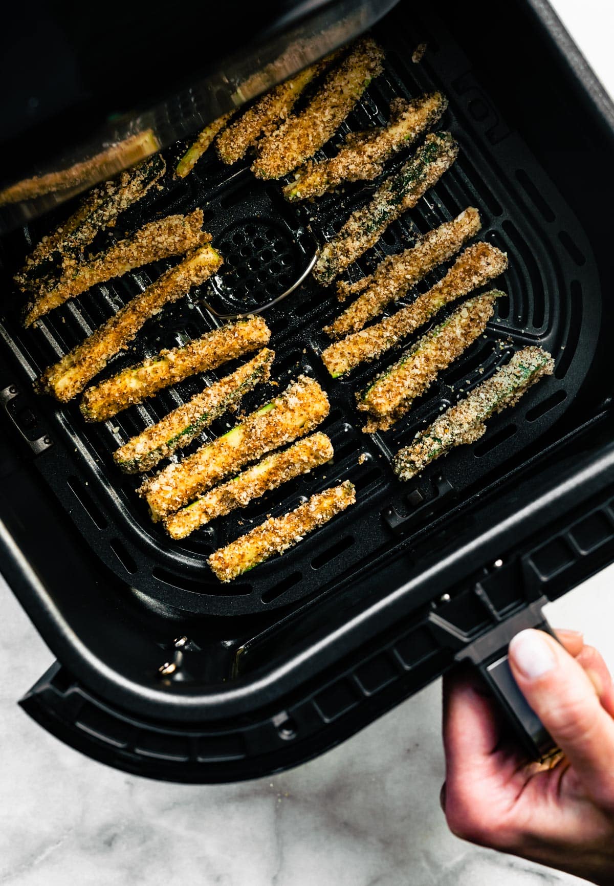 A hand holding an air fryer basket full of uncooked air fryer zucchini fries.