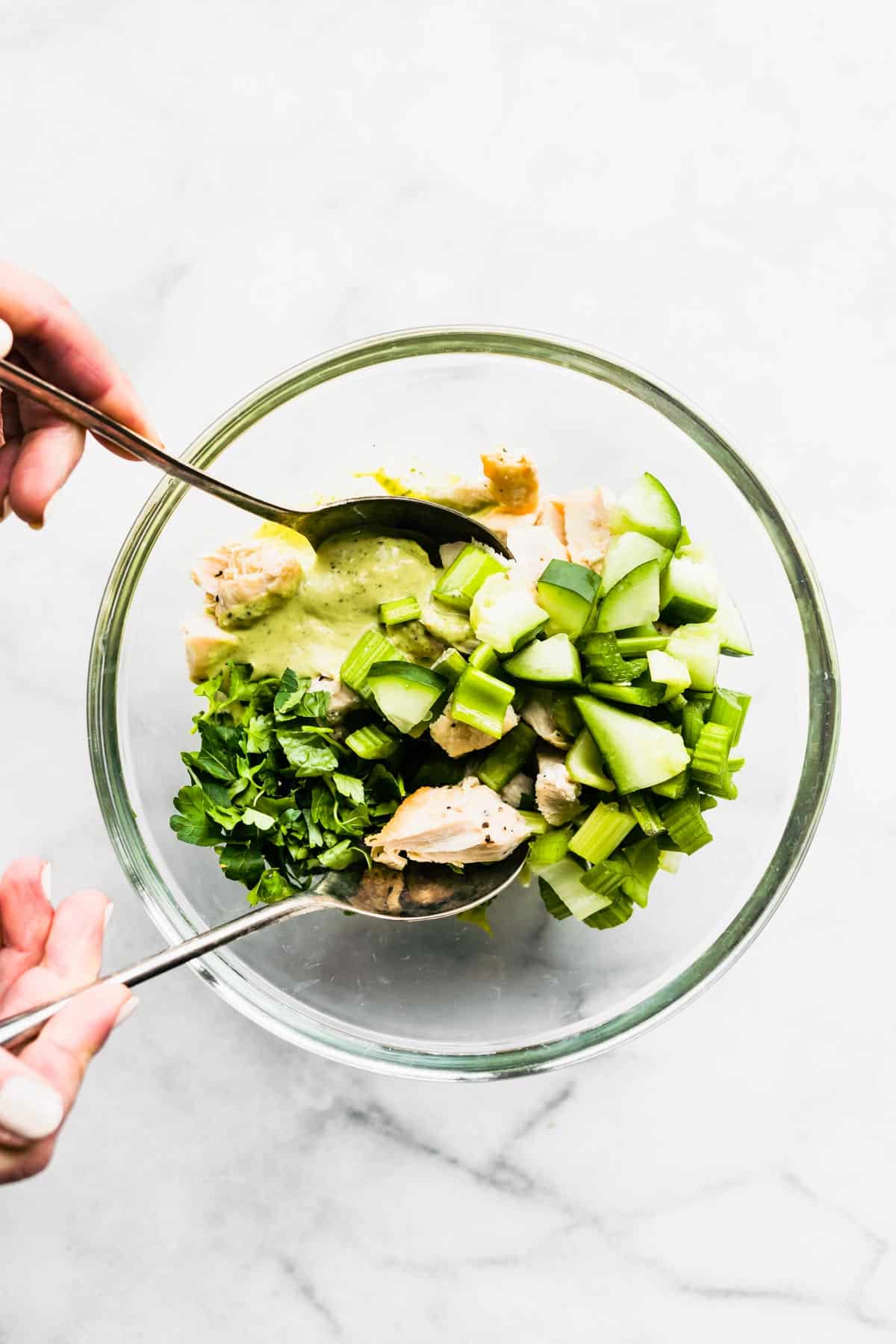 Two hands holding spoons mixing green goddess chicken salad.