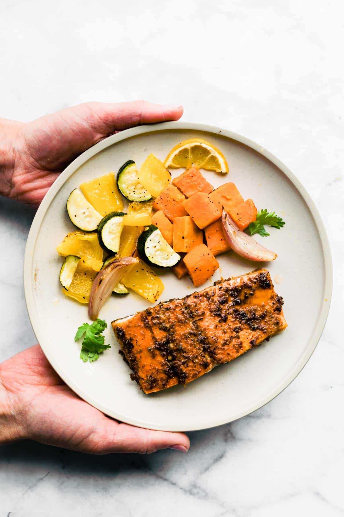 An overhead view of hands holding a white plate with jerk salmon and roasted veggies.