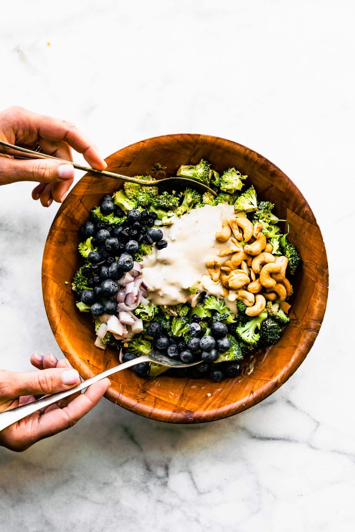 Two hands holding large spoons mixing together the ingredients of a healthy broccoli salad.