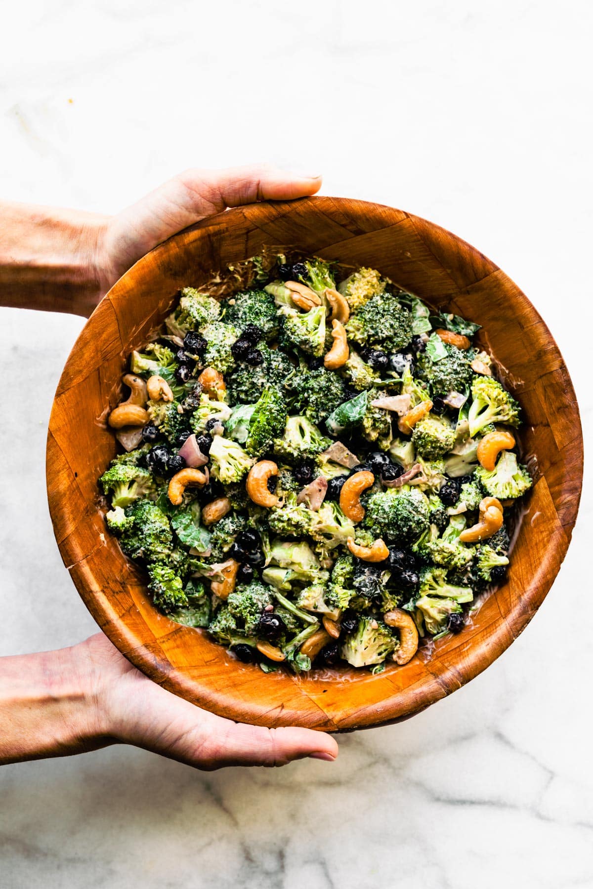 Two hands holding a wooden bowl filled with healthy broccoli salad garnished with cashews.