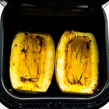 A woman's hand opening an air fryer basket with two halves of cooked spaghetti squash.