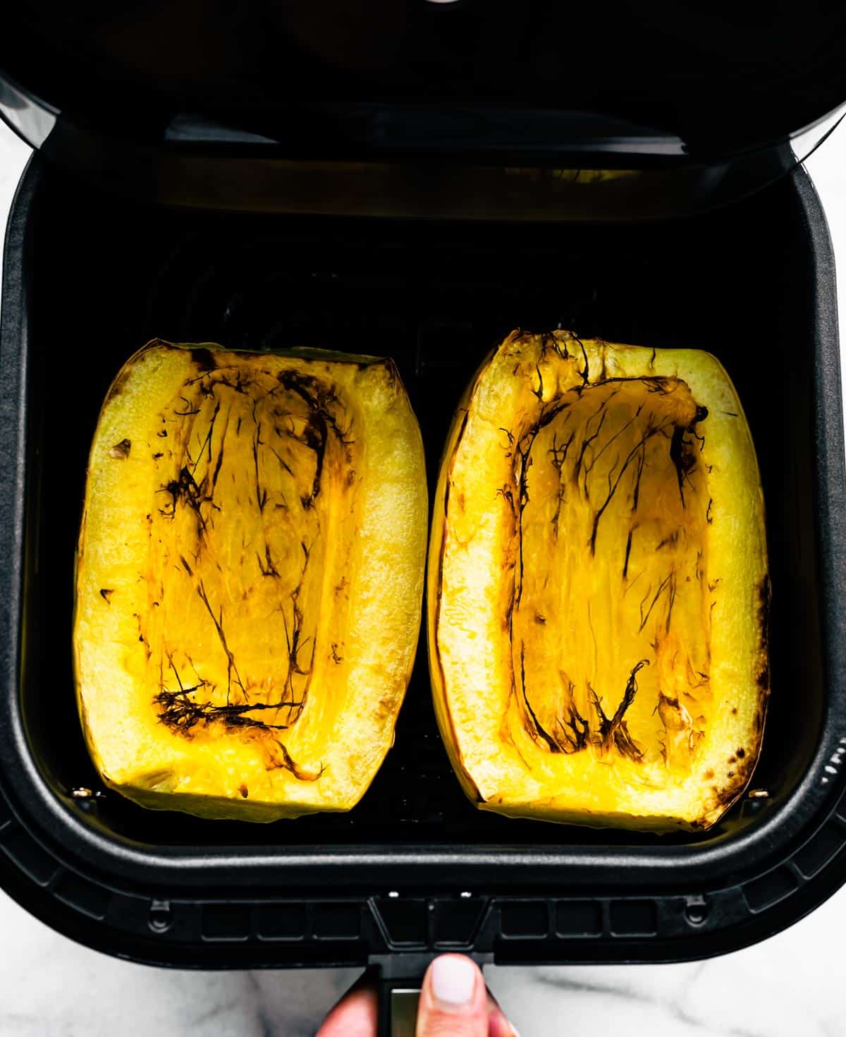 A woman's hand opening an air fryer basket with two halves of cooked spaghetti squash.