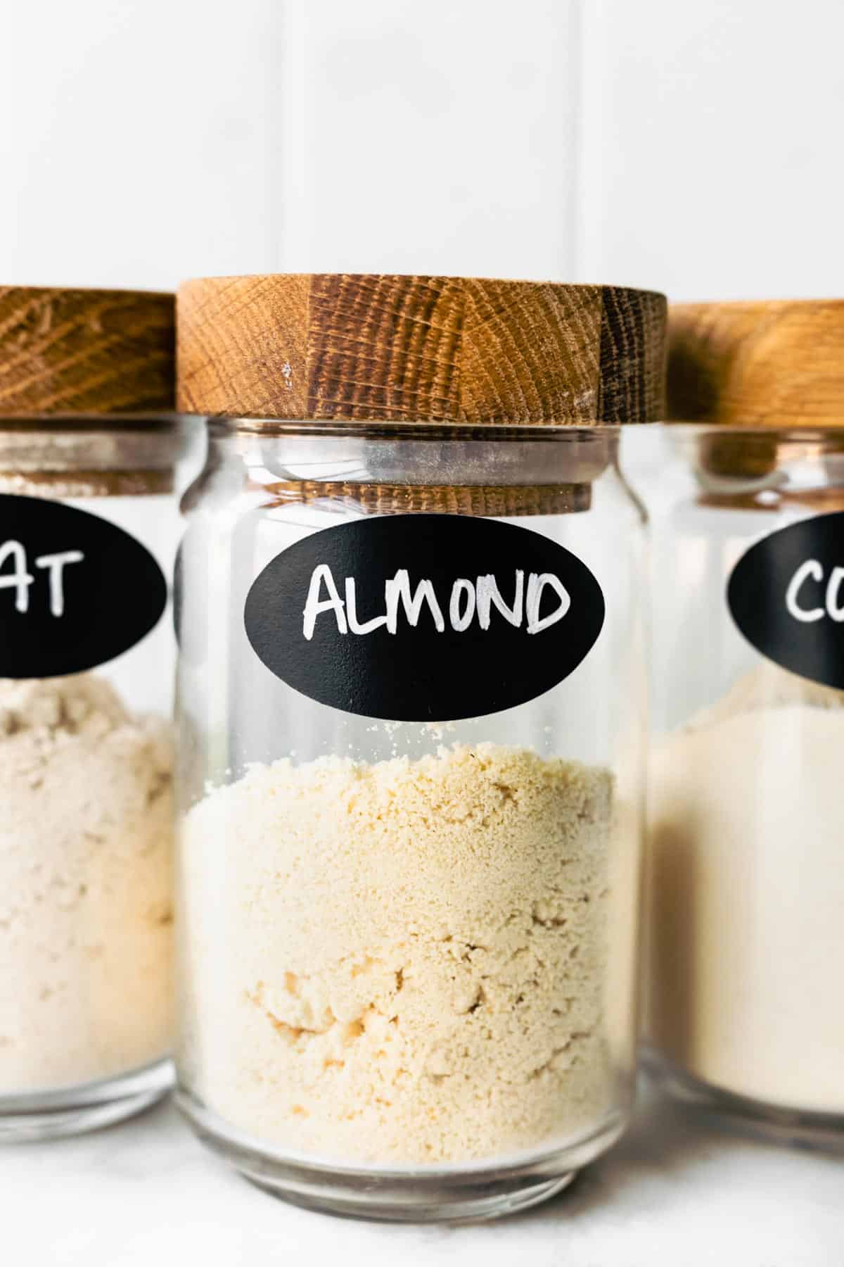 Clear glass jars of gluten free flour and almond flour with a wooden lid.