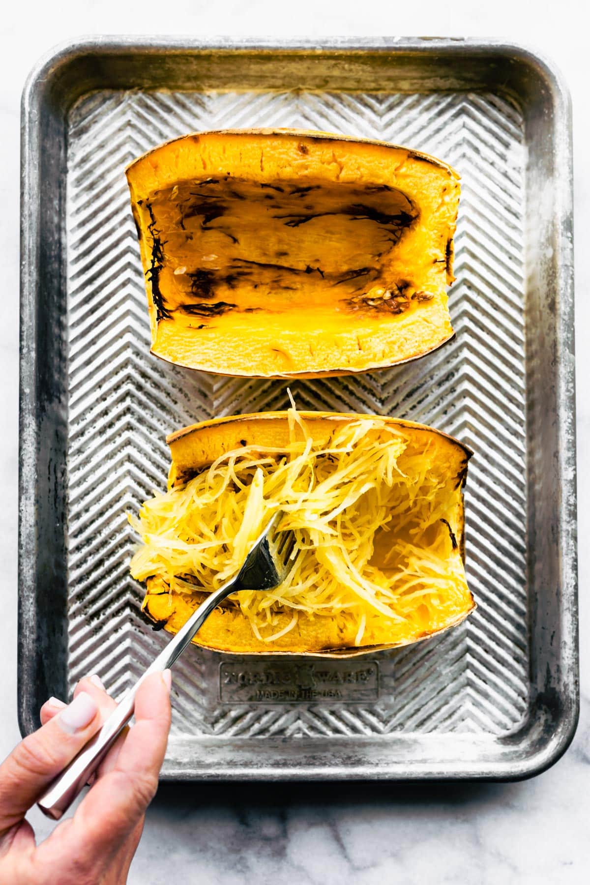 A fork peeling spaghetti squash strands from two halves of baked spaghetti squash.