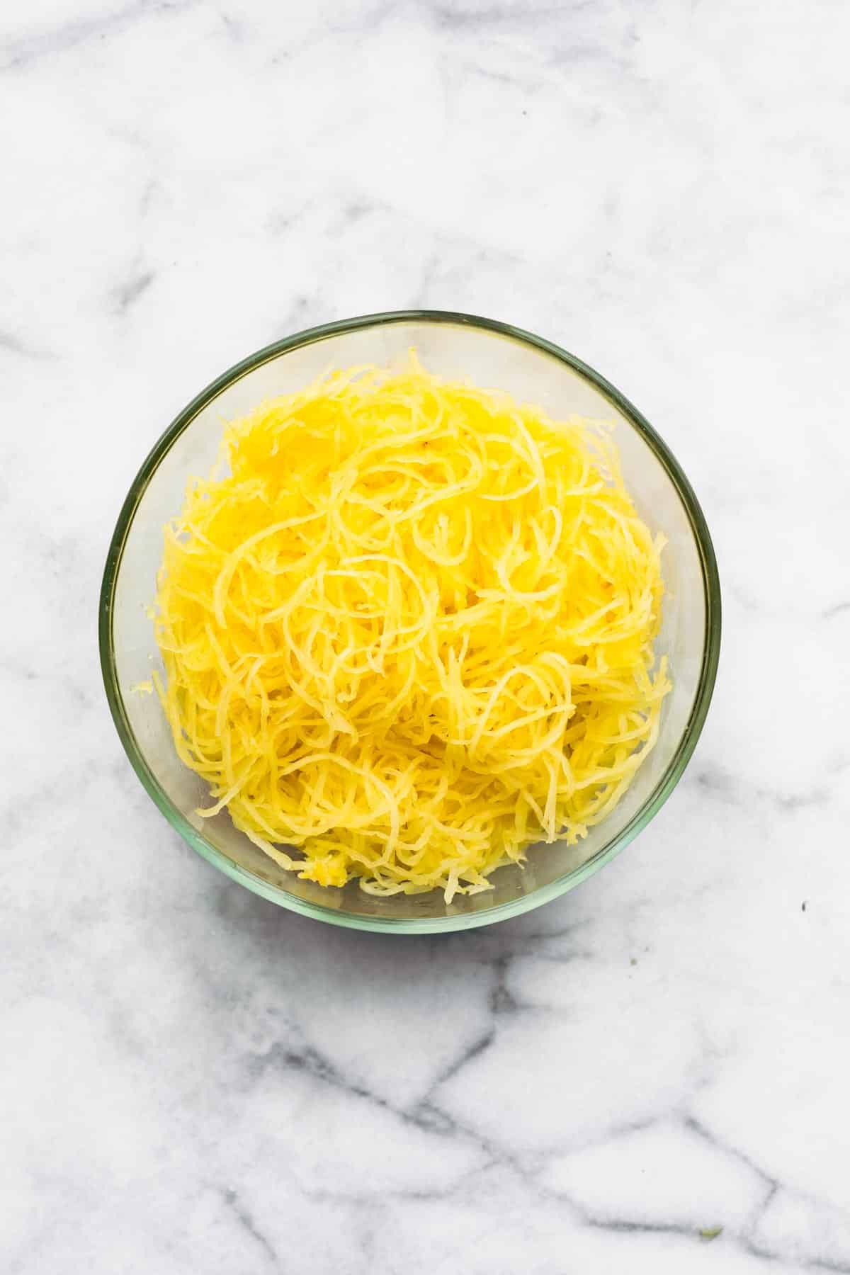 A glass bowl with spaghetti squash noodles in it on a white marble countertop.