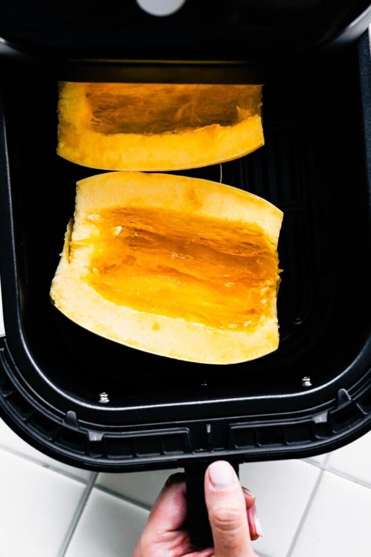A woman's hand closing an air fryer basket with two halves of raw spaghetti squash.