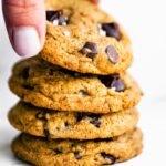 A woman's hand picking up a chocolate chip cookie from a stack of cookies.